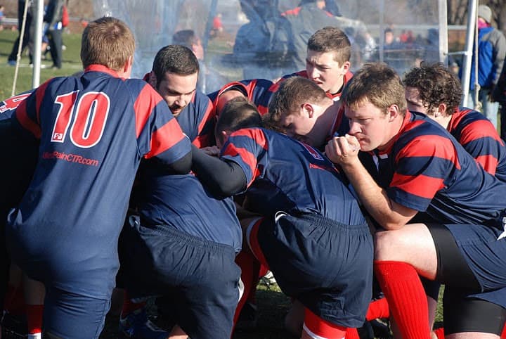 Sacred Heart Rugby men's players binding into a scrum, shoulders locked and heads down, in navy and red hooped jerseys.