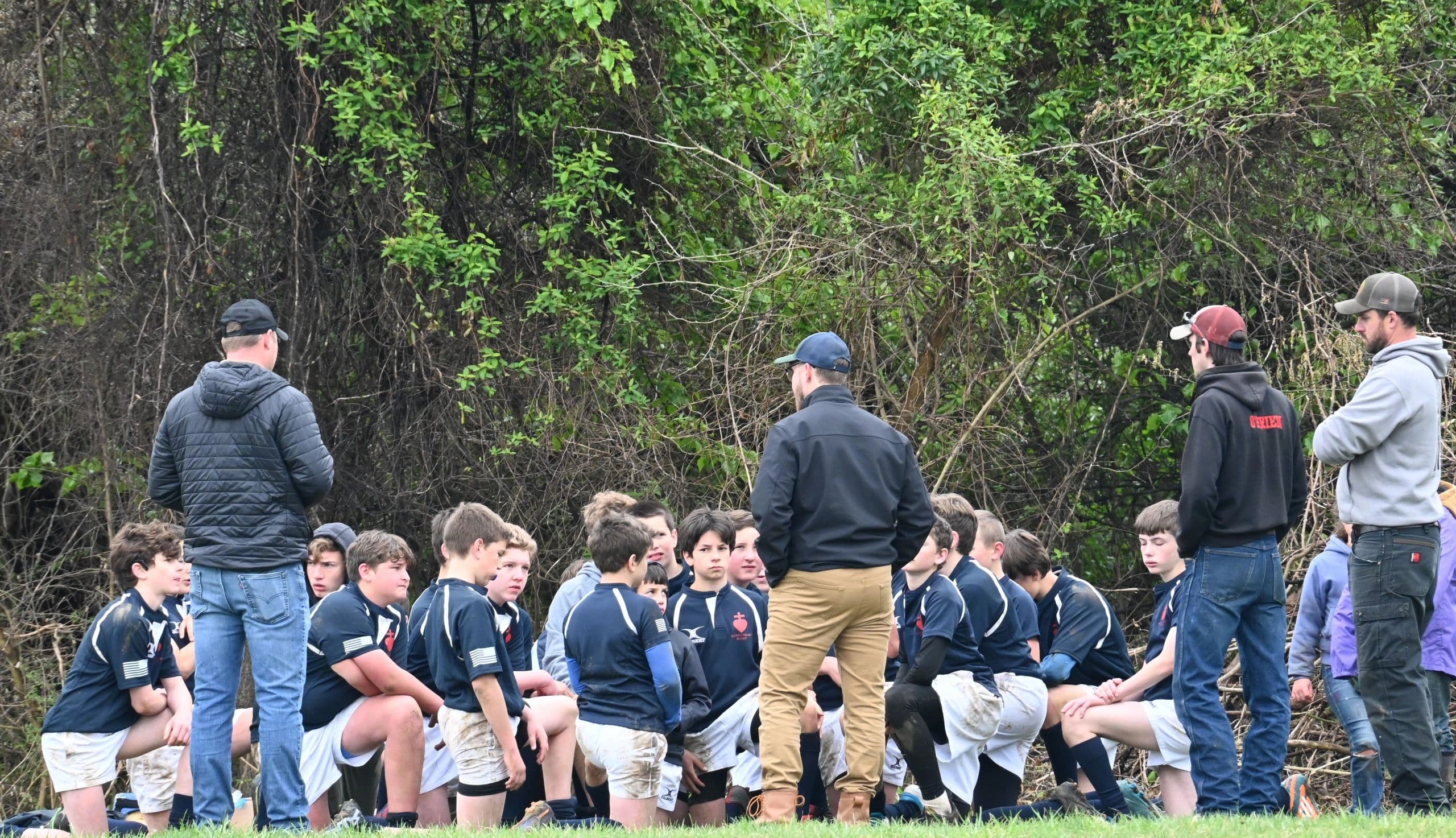 A Sacred Heart Rugby coach speaking to a group of players kneeling in their navy jerseys at the edge of the practice pitch.