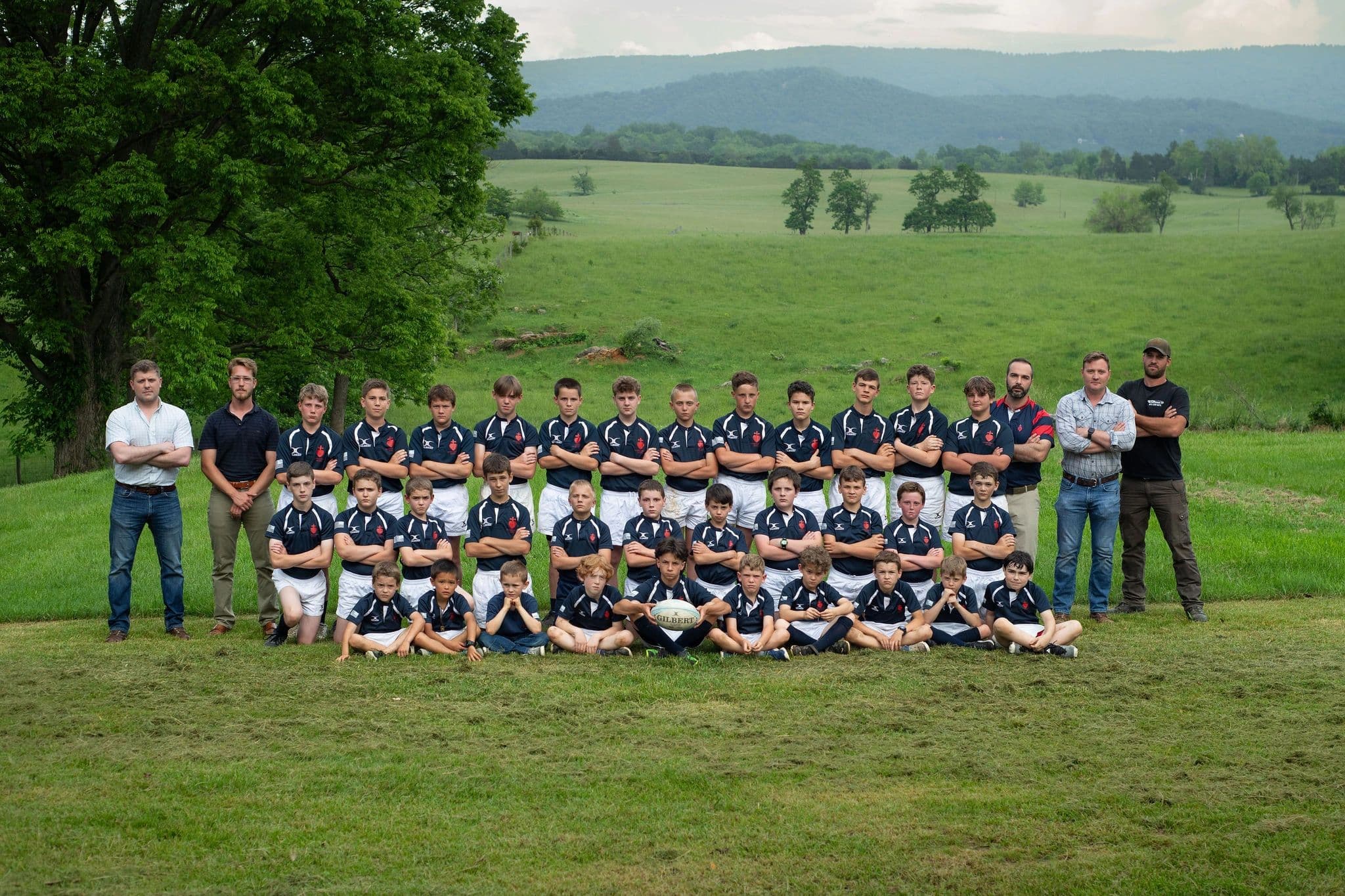 The full Sacred Heart Rugby squad and coaching staff lined up in navy jerseys on a grass pitch, with the Blue Ridge mountains behind them.